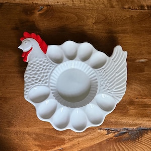May include: White ceramic egg holder in the shape of a hen. The hen has a red comb and wattle. The egg holder has spaces for 12 eggs and a center area. The egg holder is on a wooden surface.