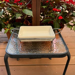 May include: A rectangular, off-white ceramic baking dish with a metal wire frame and handles. The dish is sitting on a small glass-topped table with a black metal frame. Red flowers and green plants are in the background.