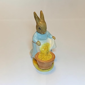 May include: A ceramic figurine of a brown rabbit wearing a blue dress, pouring yellow flower petals into a brown pot. The rabbit is standing on a yellow base.