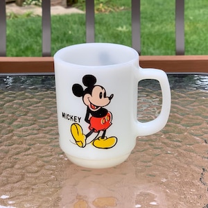 May include: White ceramic mug with a cartoon image of Mickey Mouse in red shorts and yellow shoes. The mug has a black handle and the word "Mickey" printed on the front.