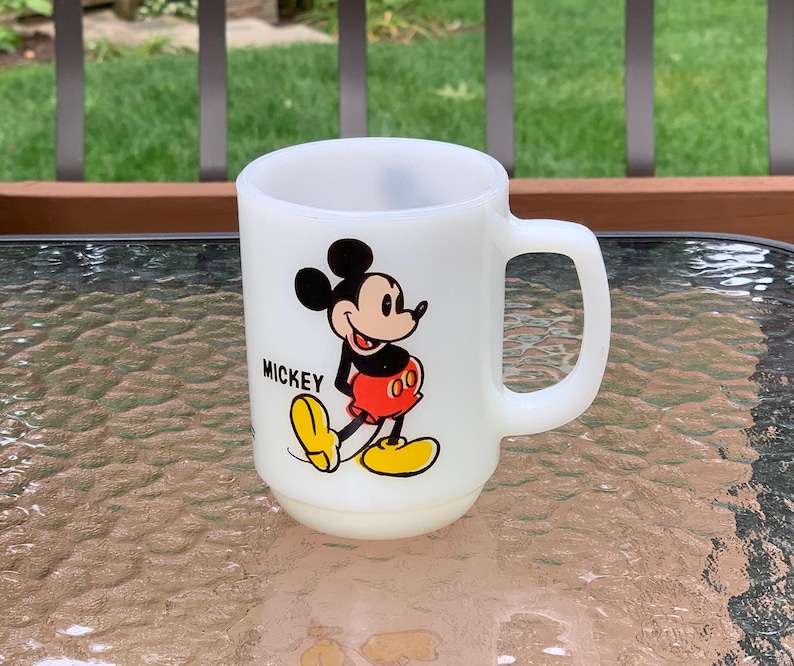May include: White ceramic mug with a cartoon image of Mickey Mouse in red shorts and yellow shoes. The mug has a black handle and the word "Mickey" printed on the front.
