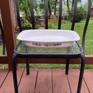 May include: A rectangular white ceramic baking dish with rounded corners and integrated handles. The exterior features a colorful decorative band of various fruits and vegetables, including red, yellow, and green produce, encircling the base.