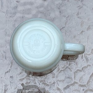 May include: White ceramic mug with a handle. The bottom of the mug has a circular logo with the text "Greengate Essen" and a small logo of a house with a heart.