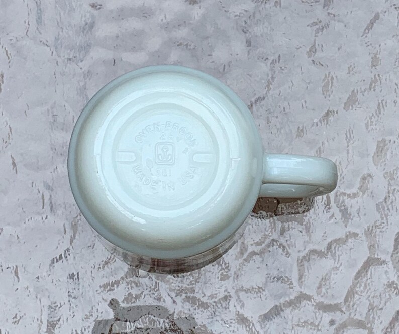 May include: White ceramic mug with a handle. The bottom of the mug has a circular logo with the text "Greengate Essen" and a small logo of a house with a heart.