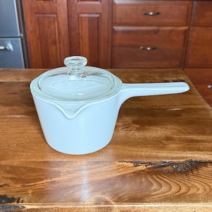 May include: A light blue saucepan with a clear glass lid and a long handle. The saucepan is sitting on a wooden surface. The kitchenware is designed for cooking and food preparation.