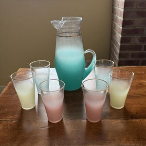 May include: A set of seven frosted glass beverageware, including a pitcher and six glasses. The pitcher is a light blue color with a gold band around the top. The glasses are in three colors: light blue, light pink, and light yellow. The glasses are all frosted and have a gold band around the top.