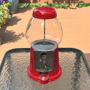 May include: A red gumball machine featuring a clear glass globe, a metallic coin mechanism with a rotating handle, and a small dispensing tray at the base. The machine has a rounded red lid and a sturdy red base, designed for holding small candies.