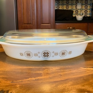 May include: White oval casserole dish with a clear glass lid. The dish features a brown and gold geometric pattern around the rim. The lid has a green tint on the edges. A vintage kitchenware item.