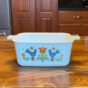 May include: A light blue rectangular casserole dish with handles. The dish features a decorative design of two blue birds, orange flowers, and green foliage. The dish is sitting on a wooden surface.