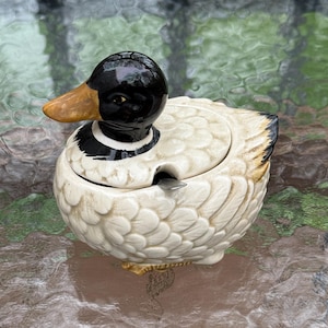 May include: A ceramic duck-shaped container with a lid and a small spoon. The duck has a black head, a yellow beak, and a white body with a textured, feather-like pattern. The container is likely for sugar or condiments.