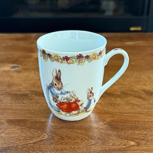 May include: White ceramic mug featuring a charming illustration of rabbits in a garden scene. The mug has a floral border and a curved handle. The design includes a rabbit wearing a blue dress and other rabbits wearing red and blue clothing.