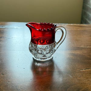 May include: A small, clear glass creamer with a red glass top. The creamer has a scalloped rim and a handle. The creamer is decorated with a pattern of raised dots.
