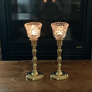 May include: Two gold candle holders with clear glass candle shades. The candle shades have a textured bubble design. The candle holders are on a wooden surface.