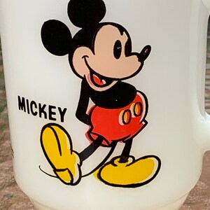 May include: White ceramic mug with a cartoon image of Mickey Mouse. Mickey is wearing red shorts with yellow buttons and yellow shoes. The mug has the word "MICKEY" printed on the front.