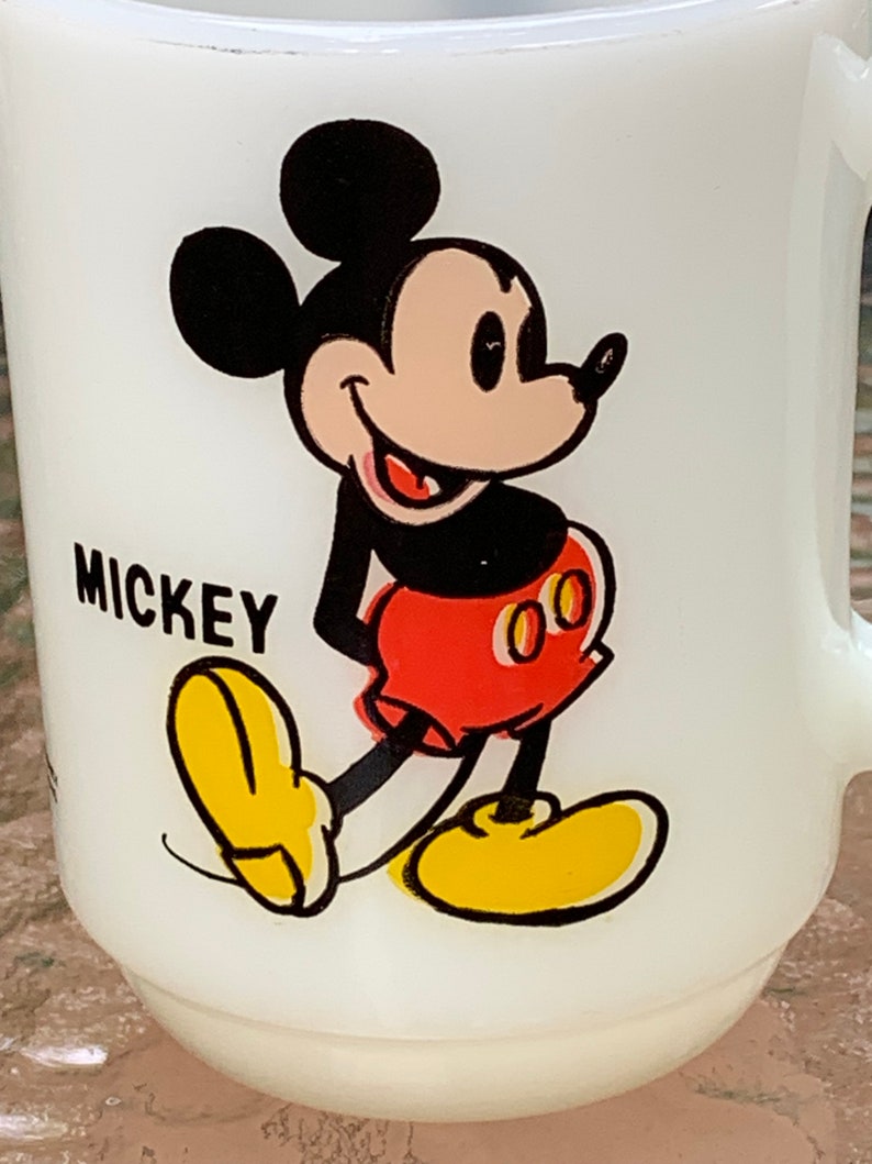 May include: White ceramic mug with a cartoon image of Mickey Mouse. Mickey is wearing red shorts with yellow buttons and yellow shoes. The mug has the word "MICKEY" printed on the front.