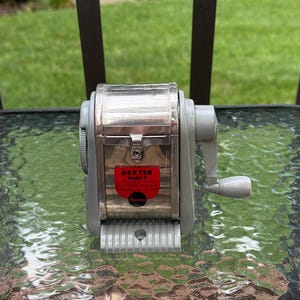May include: A vintage Dexter Model 8 pencil sharpener with a grey metal frame and a polished chrome front panel. The device features a hand crank on the right side and a red label on the front that reads Dexter Model 8 Automatic Pencil Sharpener.
