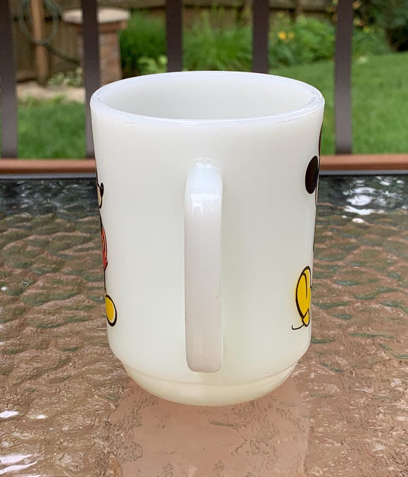 May include: White ceramic mug with a cartoon image of Mickey Mouse in red, yellow, and black. The mug has a handle.