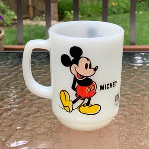 May include: White ceramic mug with a cartoon image of Mickey Mouse in red shorts and yellow shoes. The mug has the word "MICKEY" printed on the side.