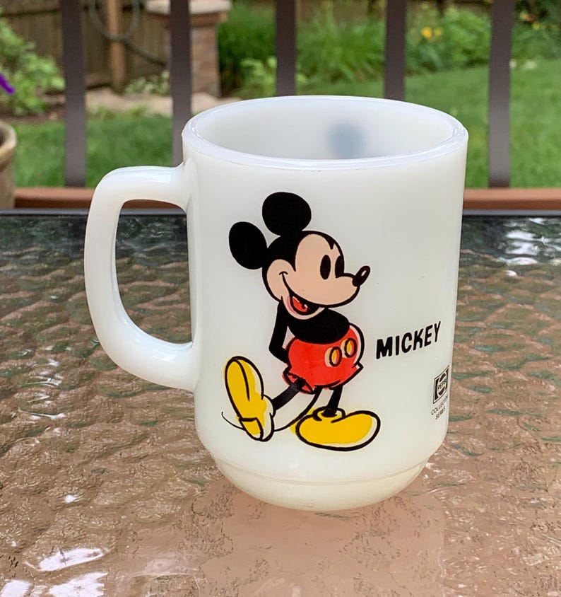 May include: White ceramic mug with a cartoon image of Mickey Mouse in red shorts and yellow shoes. The mug has the word "MICKEY" printed on the side.