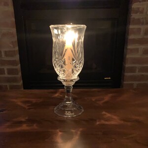 May include: A clear glass candle holder with a cut glass design. A lit candle is inside the holder.