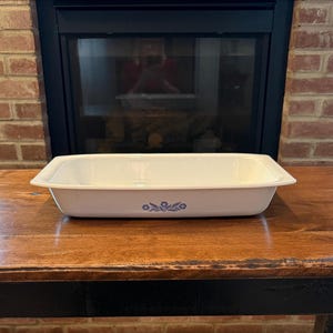 May include: A rectangular, off-white baking dish with a blue floral design on the front. The dish is sitting on a wooden table in front of a fireplace. The dish is made of a durable material.