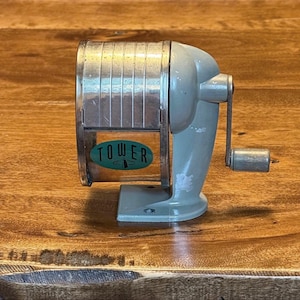 May include: A vintage manual pencil sharpener with a light gray metal body and a silver-toned cylindrical shaving receptacle. The front features a green oval label with the word TOWER printed in black. A hand crank is attached to the right side.
