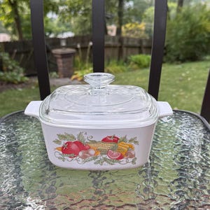 May include: White casserole dish with a clear glass lid. The dish features a colorful illustration of tomatoes, corn, and other vegetables. The dish is sitting on a glass table with a metal chair in the background.