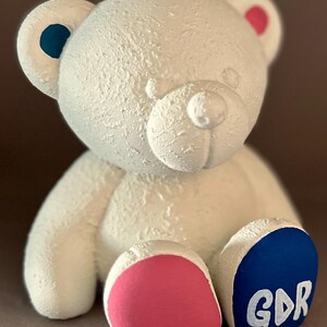 以下が含まれることがあります： テクスチャ加工が施された白いテディベアの置物。クマの耳と足には青とピンクのアクセントがあります。右足には白い文字で「GDR」と書かれています。クマは足を伸ばして座っています。