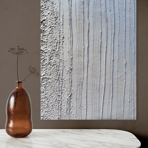 Könnte beinhalten: Ein abstraktes weißes strukturiertes Gemälde mit vertikalen und rauen Linien. Eine braune Glasvase mit getrockneten Blumen steht auf einem weißen Marmortisch. Das Kunstwerk wird vor einer braunen Wand ausgestellt.