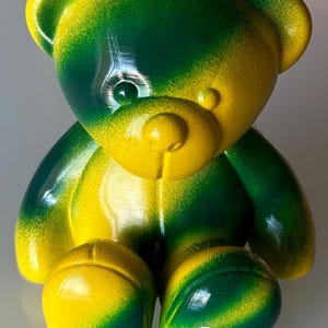 May include: A vibrant, stylized teddy bear figurine in shades of yellow and green. The bear has a glossy finish and a gradient effect, with the colors blending across its body. The letters "GDR" are visible on one foot.