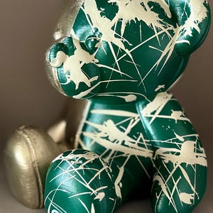 May include: A decorative bear figurine with a metallic gold head and limbs. The body is a glossy green, splattered with white paint. The bear is sitting, with the initials "GDR" visible on one foot.