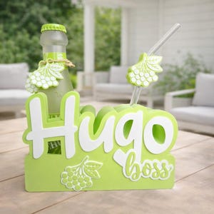 Könnte beinhalten: Ein grüner Getränkehalter mit den Worten "Hugo Boss" in Weiß. Eine klare Glasflasche mit grünem Deckel und ein Strohhalm mit Traubendesign sind zu sehen. Der Halter ist so konzipiert, dass er eine Flasche und einen Strohhalm aufnehmen kann.