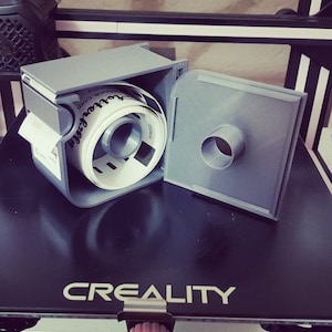 May include: A gray 3D printed label dispenser with a roll of white labels inside. The label roll has black text that reads "Got Your Back" and "shopify.com". The dispenser has a hinged lid that is open, revealing the label roll.