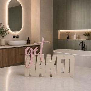Könnte beinhalten: Dekoratives Schild in einem Badezimmer. Das Schild zeigt "get NAKED" in einer Kombination aus Rosa und Beige. Das Badezimmer hat ein modernes Design mit Badewanne, Waschbecken und Spiegel.