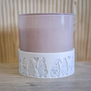 Puede incluir: Una vela en un recipiente de vidrio morado claro, colocada sobre una base de cerámica blanca. La base presenta un patrón repetido de figuras de gnomos sosteniendo corazones. El fondo es una superficie de madera clara.