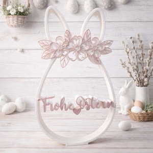 Puede incluir: Decoración blanca con forma de conejito de Pascua con una corona floral y las palabras "Frohe Ostern" en rosa. La decoración tiene un diseño recortado y está colocada sobre una superficie de madera blanca. Decoración temática de Pascua.