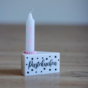 Könnte beinhalten: Eine weiße Kerze mit rosa Schriftzug "Alles Gute" steht in einem weißen kuchenförmigen Halter mit schwarzen Tupfen und dem Wort "Pustekuchen". Der Kerzenhalter hat eine rosa Perlenborte.