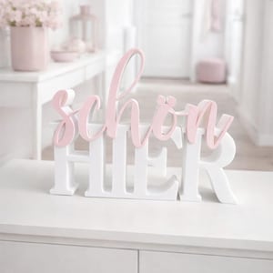 May include: A white wooden sign with the word "HIER" in capital letters and the word "schön" in pink cursive letters. The sign is displayed on a white surface, with a blurred background of a room decorated in shades of pink and white.