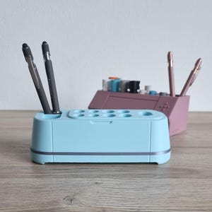 Może przedstawiać: Dwa pojemniki na długopisy na zdjęciu. Przedni jest jasnoniebieski z szarym obramowaniem, trzymający dwa długopisy. Drugi pojemnik na długopisy jest różowy i mieści dwa długopisy. Tło to jasna ściana i drewniana powierzchnia.