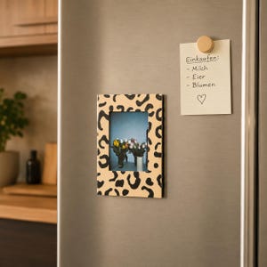 May include: A rectangular picture frame with a leopard print pattern is attached to a refrigerator. The frame holds a photo of flowers in vases. A wooden magnet holds a shopping list that reads "Einkaufen: - Milch - Eier - Blumen" with a heart symbol.