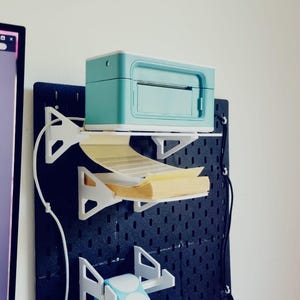 May include: A teal label printer is mounted on a black pegboard wall with a white shelf. A roll of white round labels is mounted on a white shelf below the printer. The printer is plugged into an outlet.