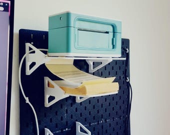 May include: A teal label printer is mounted on a black pegboard wall with a white shelf. A roll of white round labels is mounted on a white shelf below the printer. The printer is plugged into an outlet.
