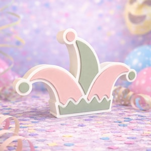 May include: A decorative jester hat in pastel colors. The hat features a white base with pink and green accents. The hat has a scalloped edge and is decorated with small, round details. The background includes confetti and balloons.