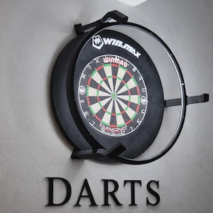 Może przedstawiać: Czarna tarcza do darta z przezroczystą obudową, zamontowana na ścianie. Tarcza do darta ma klasyczny design z czerwonymi, zielonymi i białymi segmentami. Słowo "Darts" jest wyświetlane poniżej.