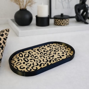 May include: An oval decorative tray with a black rim and a beige surface featuring a black leopard print pattern. The tray is placed on a white surface, with other decorative items in the background.