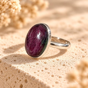 Può includere: Un anello in argento con una grande pietra preziosa lucida di forma ovale. La pietra presenta tonalità profonde di magenta scuro e viola con sottili inclusioni nere. La fascia in metallo è liscia, lucida e riflettente, e sostiene la pietra in una semplice montatura a castone.
