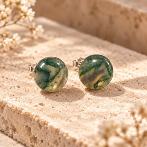 Può includere: Un paio di orecchini a lobo rotondi con pietre di agata muschiata lucide che presentano inclusioni verde scuro, traslucide e bianco nuvoloso. Le pietre sono montate su perni in metallo color argento con chiusure a farfalla sul retro.
