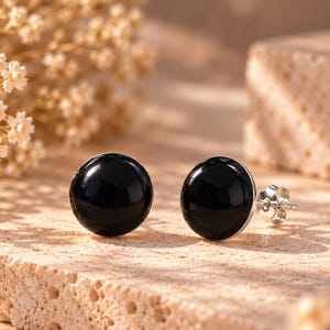 Può includere: Un paio di orecchini a lobo rotondi con pietre nere lisce e lucide incastonate in metallo color argento. Ogni orecchino ha una classica forma circolare con una finitura lucida, che mette in risalto un colore nero intenso e profondo.