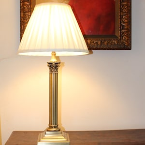 Puede incluir: Una lámpara de mesa de latón con una pantalla blanca plisada. La lámpara tiene una base de columna con una base cuadrada y una parte superior decorativa. La lámpara está sobre una mesa de madera oscura, con una pintura enmarcada en el fondo.