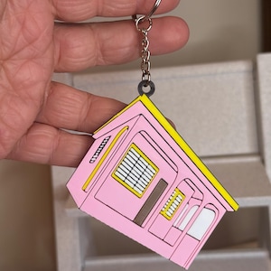 May include: A pink house-shaped keychain with yellow trim and a silver key ring. The keychain features a detailed design of a house with windows and a door.